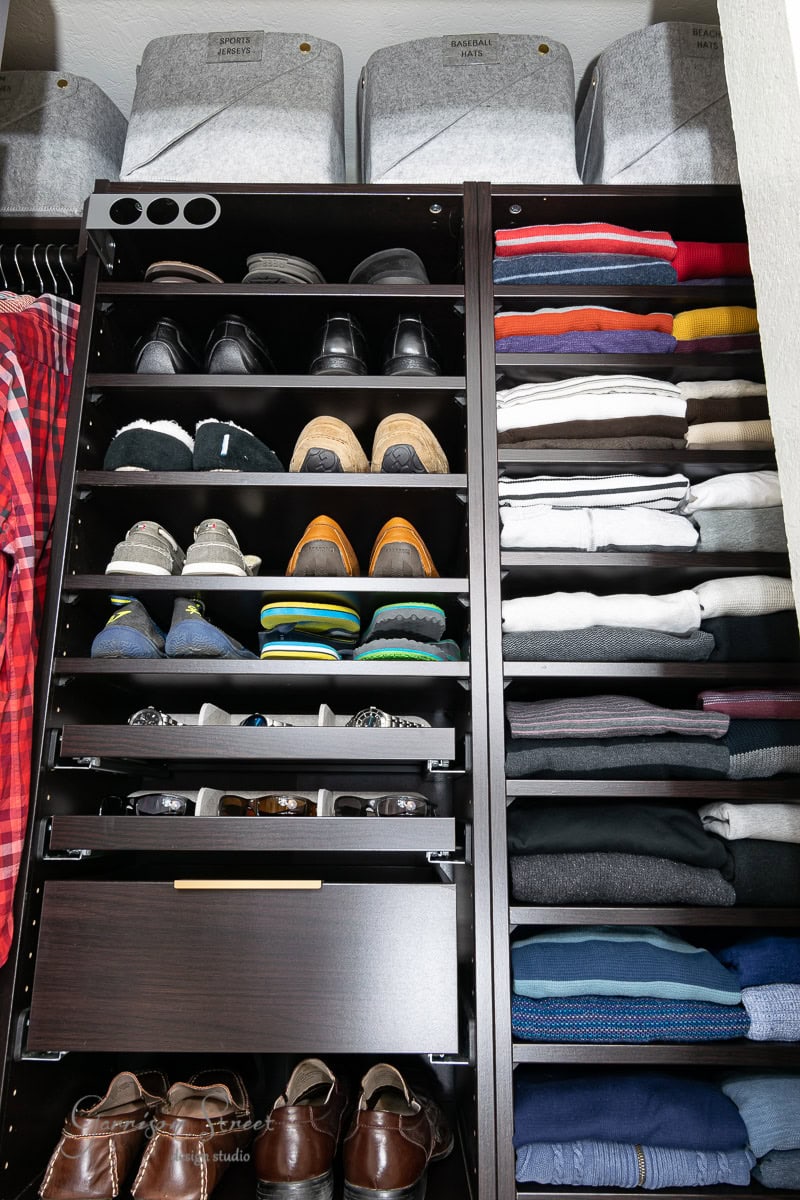 A neatly organized IKEA Pax reach-in closet features shelves of folded clothes arranged by color on the right. On the left, several pairs of shoes and accessories are displayed. Grey storage bins labeled "SPORTS" sit on the top shelf, while brown shoes rest neatly on the floor.
