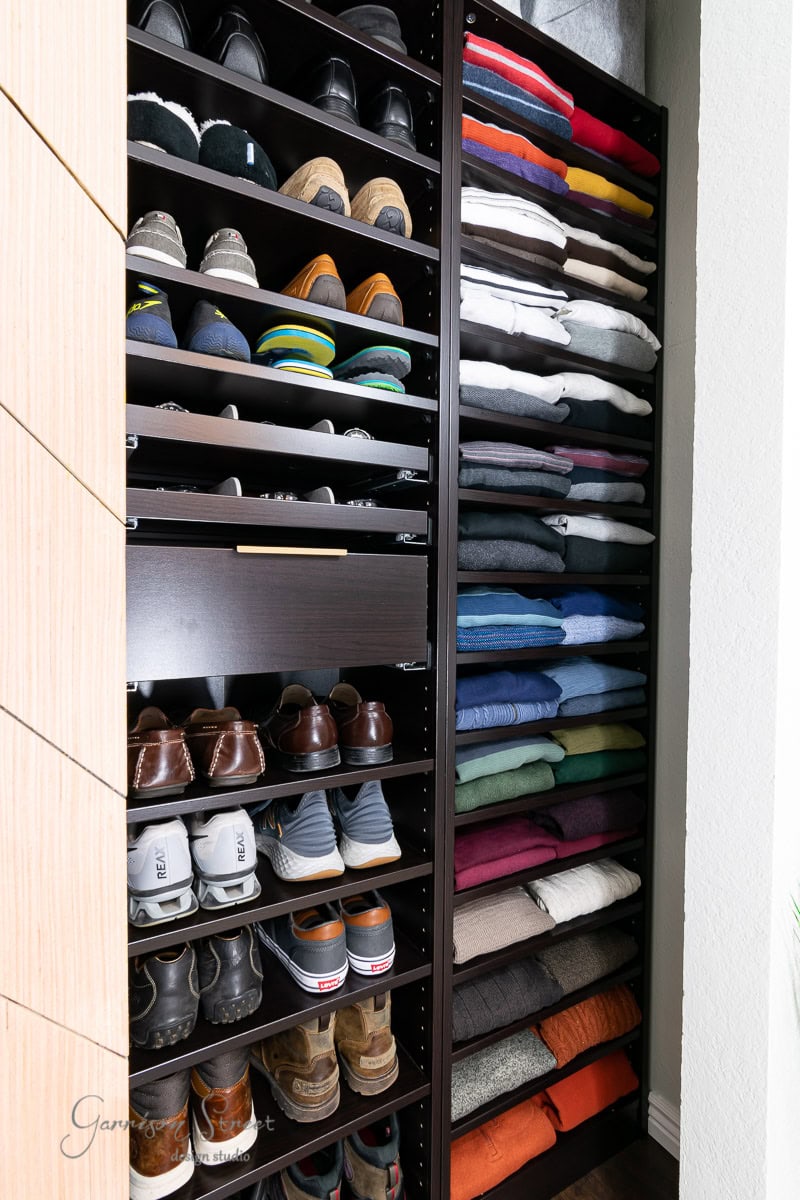 A neatly organized IKEA Pax reach-in closet with shelves full of folded clothes in various colors on the right and several pairs of shoes, including sneakers and dress shoes, on the left. The closet features dark wood shelving.
