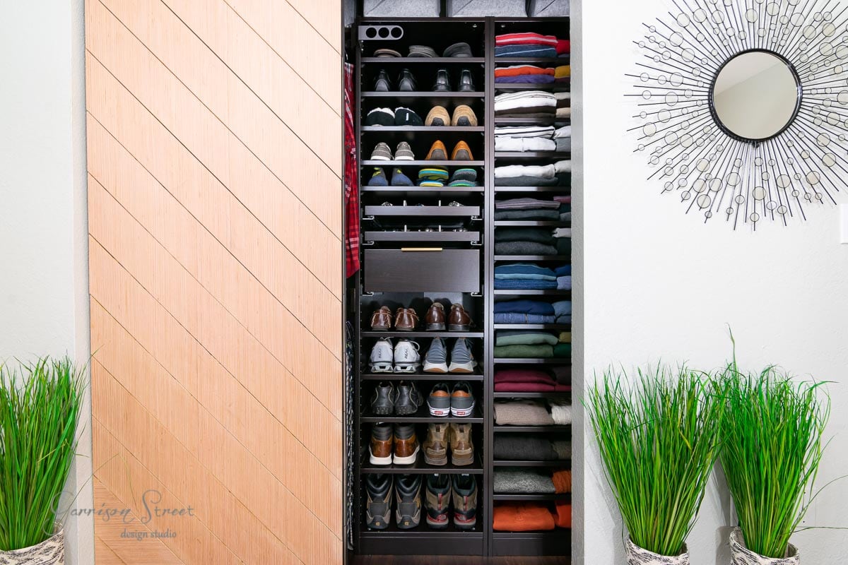 A neatly arranged IKEA Pax closet features shelves with folded clothes, organized shoes, and hats. The reach-in closet boasts a sliding wooden door. To the right, there's a round decorative mirror on the wall, complemented by green plants on either side.