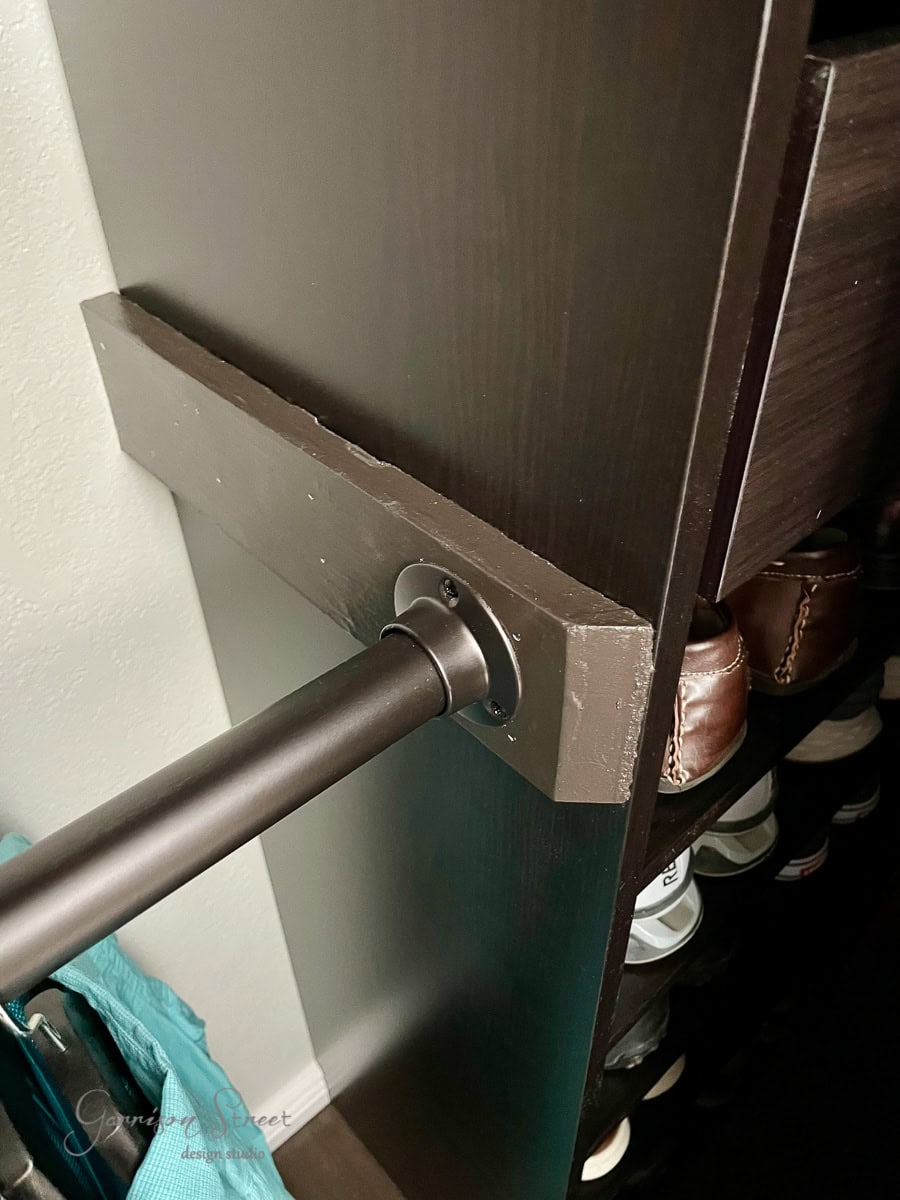 A close-up of a clothing bar installed in a dark wooden IKEA Pax closet. The bar is attached to a rectangular bracket on the side panel, perfect for maximizing space in your reach-in closet. Several shelves with neatly arranged shoes are visible in the background, completing this efficient design.