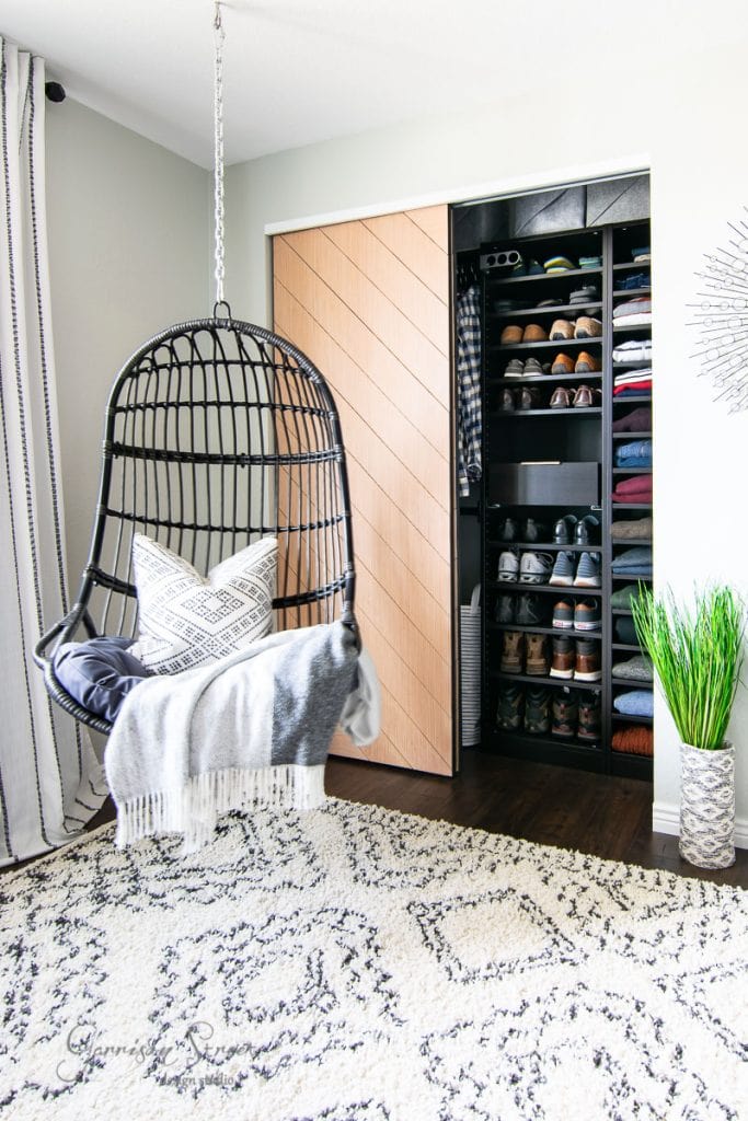 Home A modern room with a hanging wicker chair, draped with a gray blanket and patterned pillow. The reach-in closet features neatly arranged shoes and clothes. Decor includes an IKEA Pax-inspired rug, curtains, and a tall plant in a decorative vase.