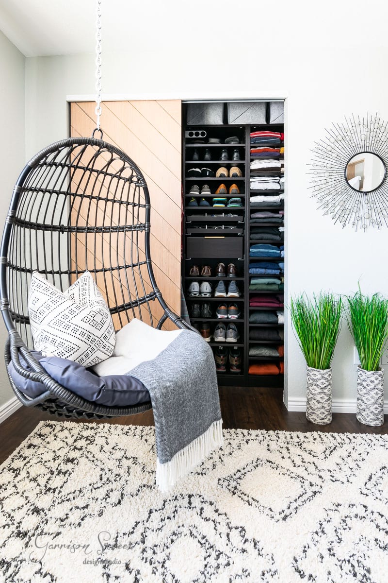 A cozy room features a hanging rattan chair with a geometric pillow and gray throw. An open IKEA Pax closet displays shoes and folded clothes. The space is decorated with a patterned rug, two potted plants, and a round mirror on the wall.
