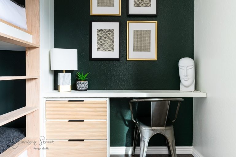 A modern study nook with a sleek metal chair and a white desk against a dark green wall exudes an expensive charm. The desk features an abstract face sculpture, a potted plant, and a contemporary lamp. Above, three framed abstract artworks are arranged vertically—perfect IKEA hacks inspiration.