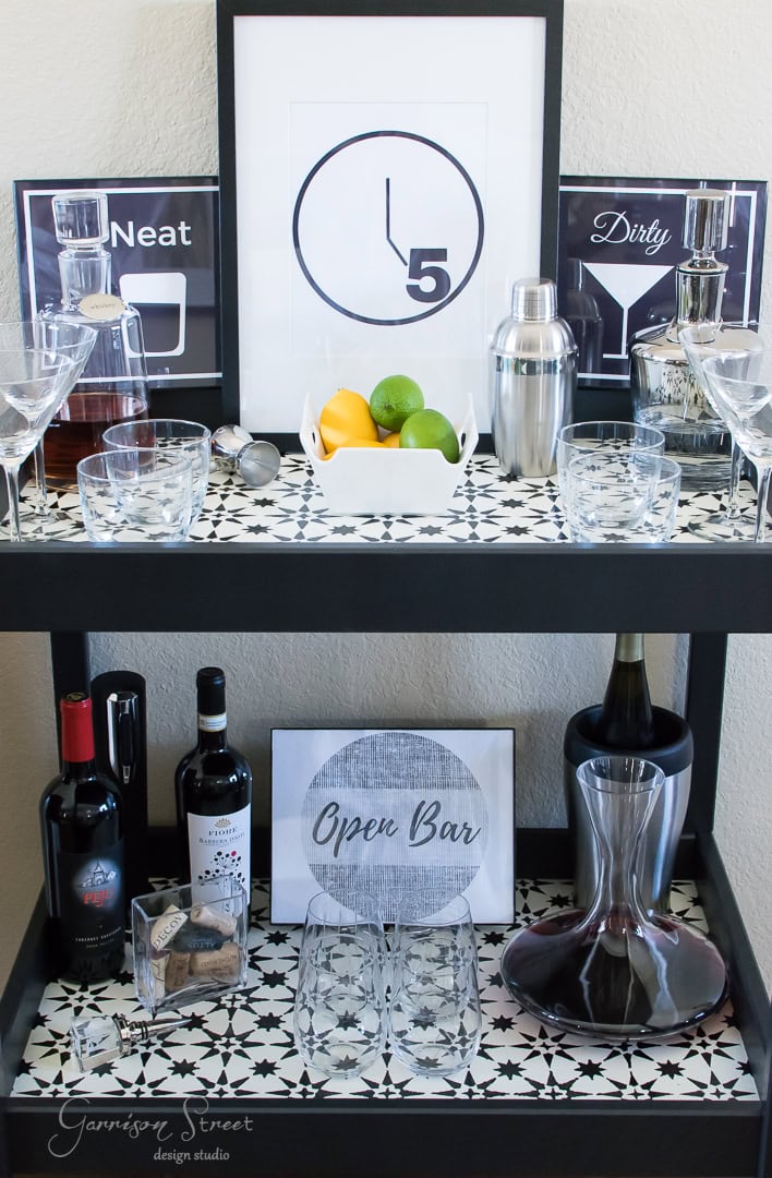 A stylish bar cart with two shelves featuring expensive liquor bottles, a decanter, cocktail shaker, wine glasses, and citrus fruits. Framed wall art with cocktail graphics and "Open Bar" sign adorn the setup. Black and white patterned surface offers chic Ikea hacks inspiration.