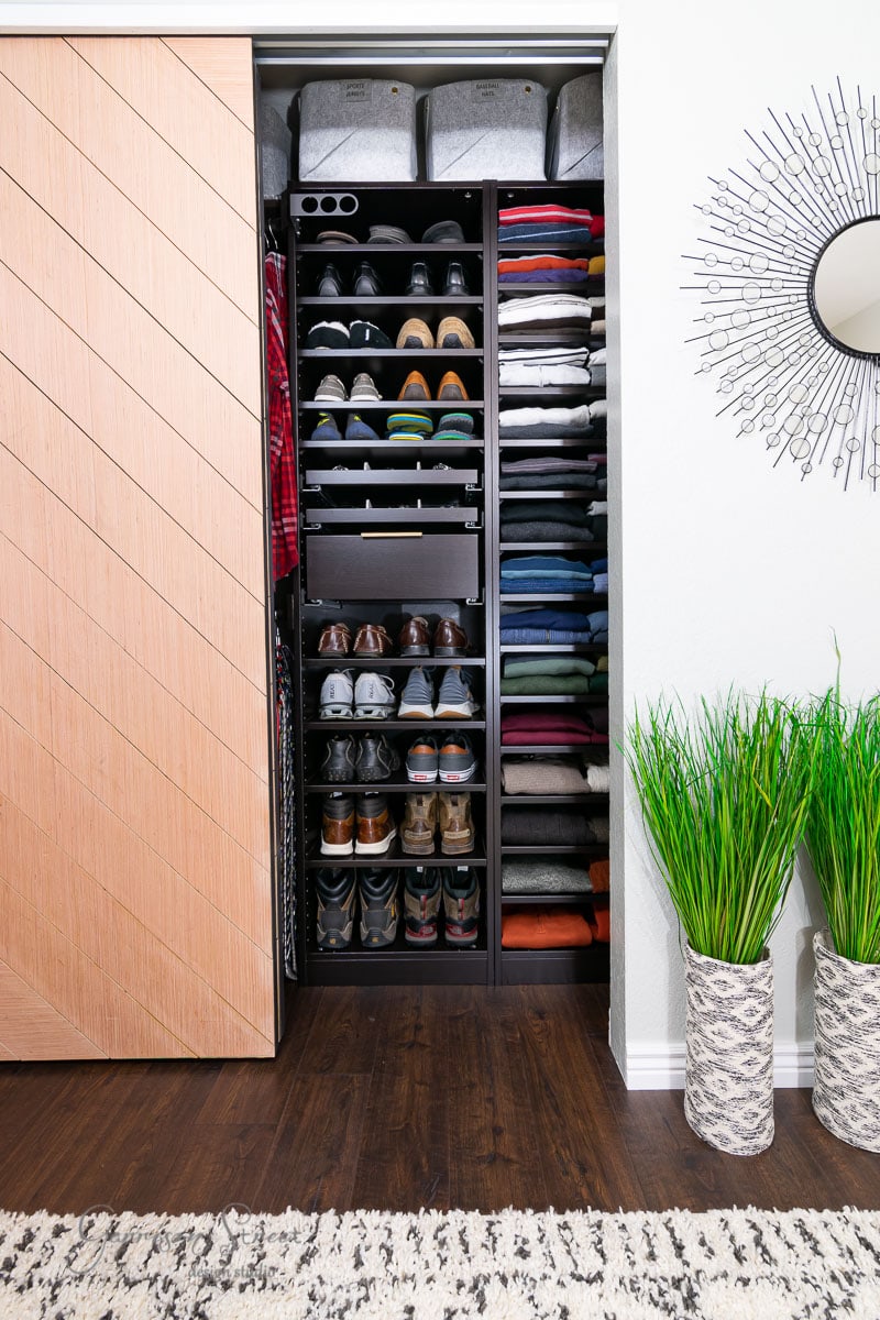 A neatly organized closet showcases shelves filled with folded clothes, stacked shoes, and bins on the top shelf. A sliding wooden door partially covers the closet. Two green plants in white pots sit on the floor, incorporating clever IKEA hacks that give it a more expensive look.