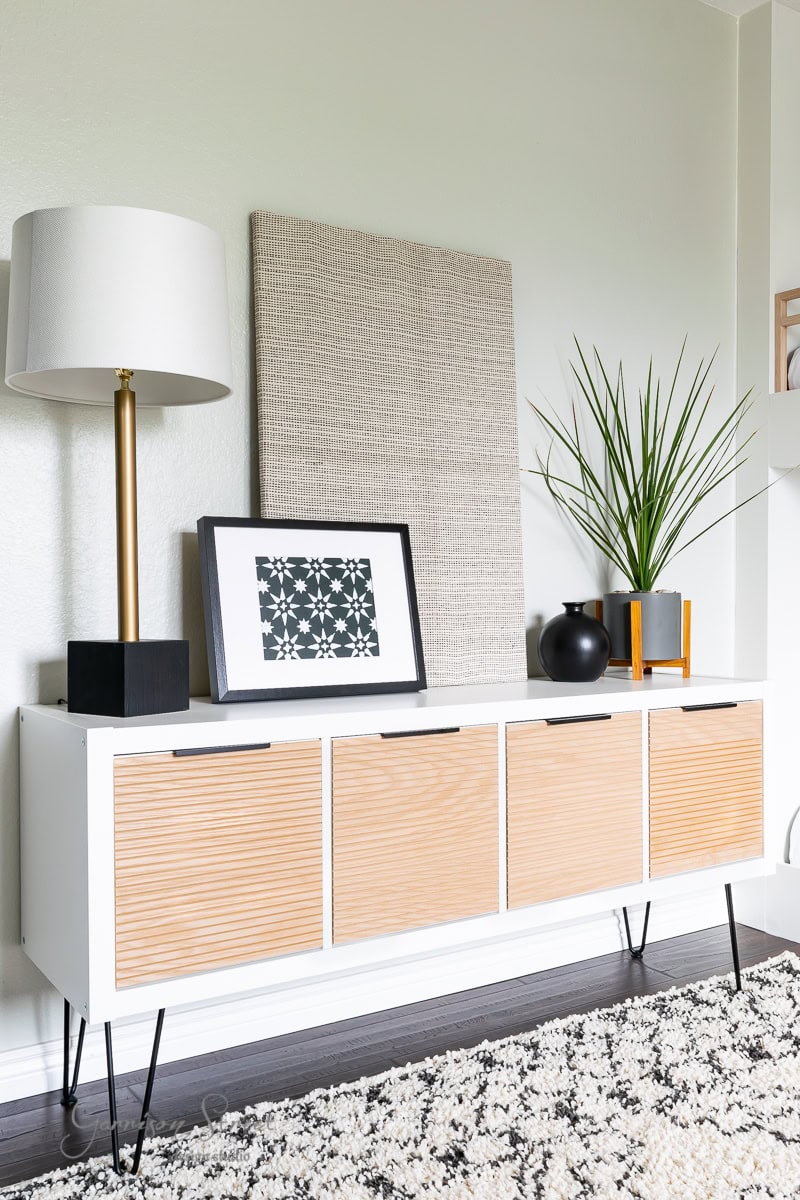 A modern living room corner showcases an expensive white sideboard with wooden front panels. Atop, a lamp with a white shade, framed art, a small plant in a black vase, and decorative items elevate the look. A textured wall art piece leans elegantly against the light-colored wall—perfect for chic IKEA hacks.