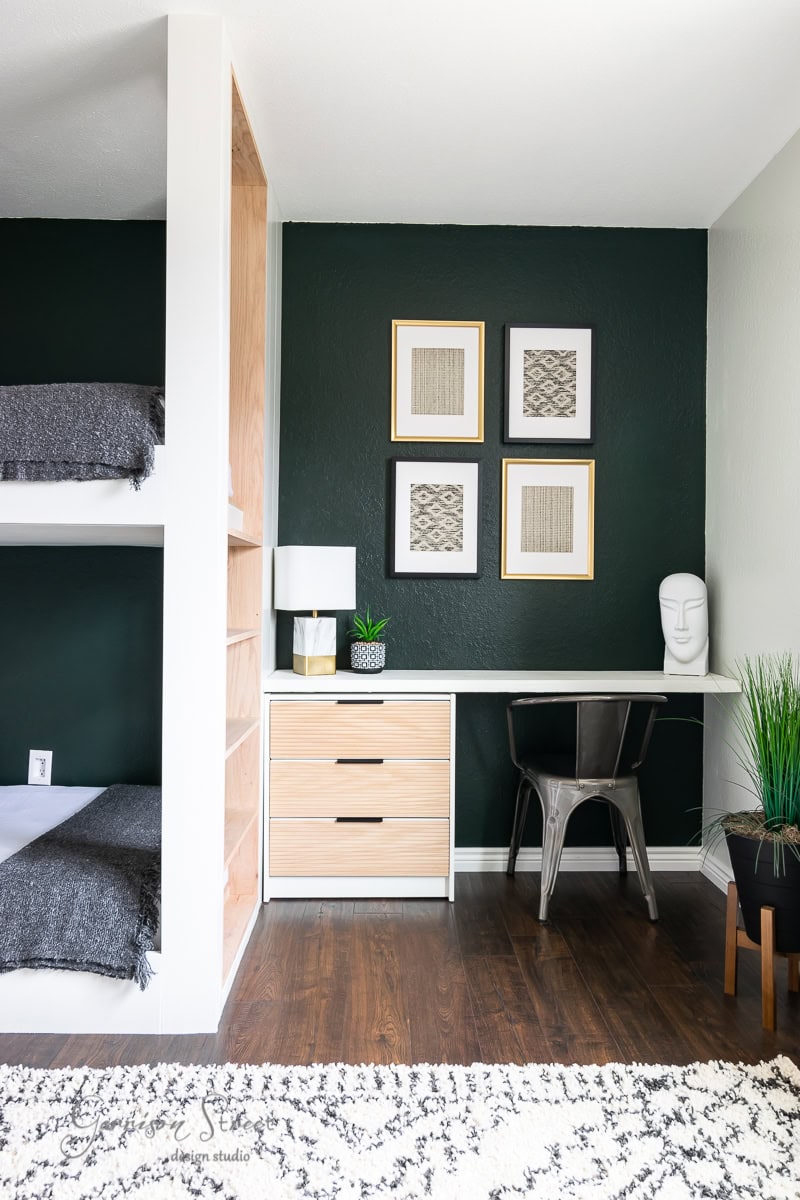 Modern room with dark green walls featuring a stylish bunk bed, desk with two drawers, and a chair. Decor includes an elegant table lamp, plant, abstract framed art, and a head sculpture. Clever IKEA hacks elevate the space's aesthetic alongside hardwood flooring and a gray rug.