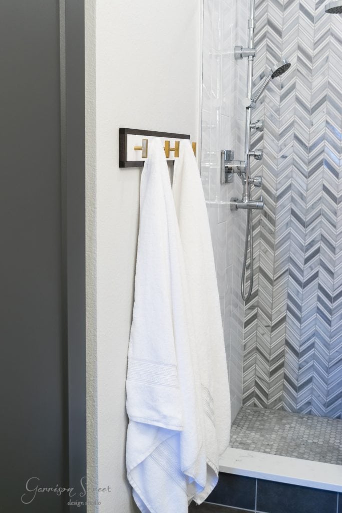 Home A modern bathroom features a sleek shower with chevron-patterned tiles in shades of gray, exuding an expensive feel. Two white towels hang neatly on a black and gold towel rack, cleverly mounted beside the shower, reminiscent of ingenious Ikea hacks for maximizing style.