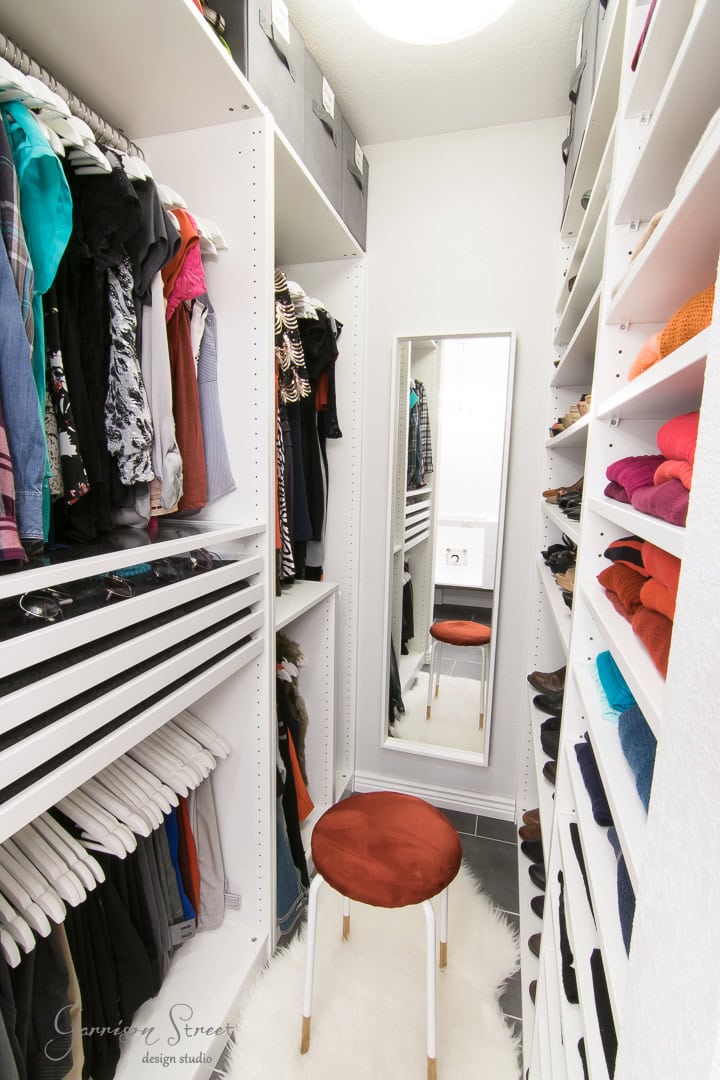 A well-organized walk-in closet with white shelves displaying neatly folded clothes, hanging garments, and shoes. A full-length mirror on the back wall adds an expensive touch, while a round stool with an orange cushion sits on a white rug in the center—a nod to clever IKEA hacks.