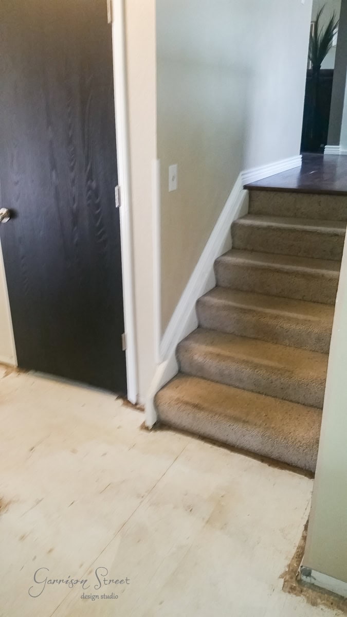 Partially renovated hallway with bare subflooring next to a staircase covered in beige carpet. A black door is on the left, and walls are painted light beige. A partial view of decor can be seen at the top of the stairs.