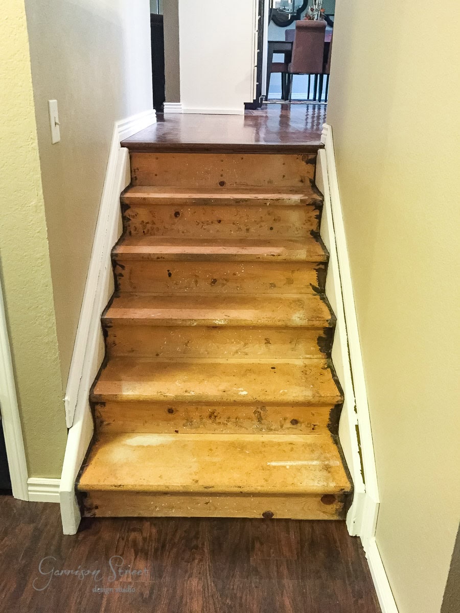 A short staircase with exposed wooden steps, showing signs of wear and stains, is nestled between two white walls. It ascends to a higher level, with a view of a dining table beyond.