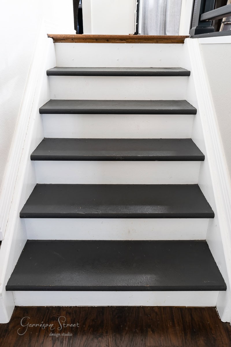 A staircase with dark gray steps and white risers shows off a sleek remodel. The wooden handrail is barely visible at the top. In this interior setting with hardwood floors, subtle branding text appears in the bottom left corner.