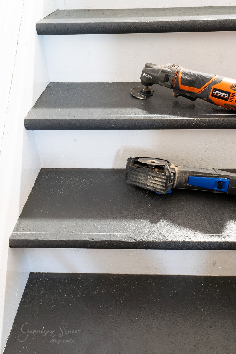 A set of indoor stairs with dark treads and light risers. An orange and black power tool sits on one step beside a tool with a blue handle. The staircase is slightly worn with specks of dirt.