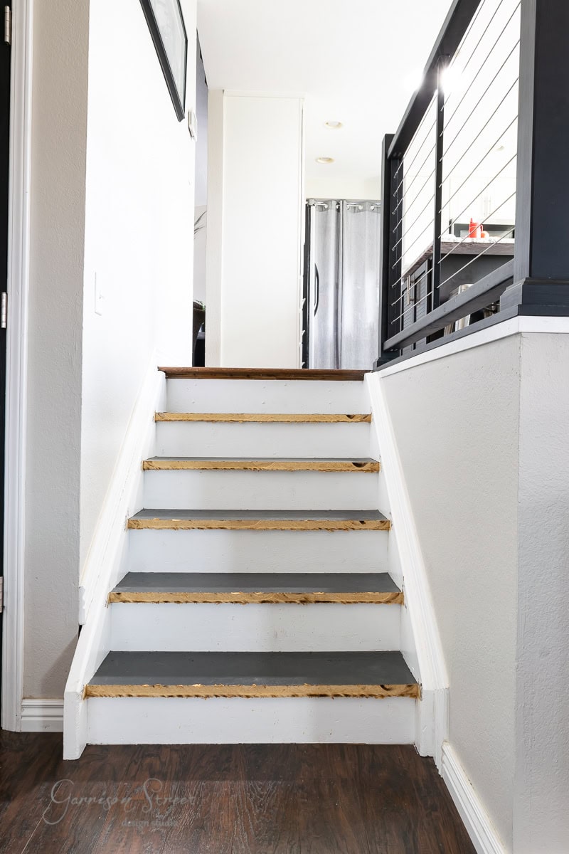 A small indoor staircase features a wooden floor, white walls, and gray steps. The black railing has horizontal metal bars. A refrigerator is visible at the top near an open doorway.