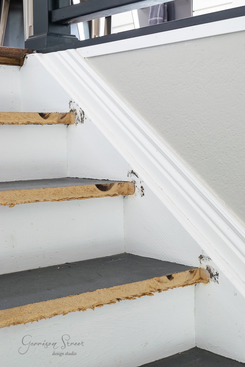 A close-up of a partially remodeled staircase with exposed edges and scuffs. The steps are painted gray, showing visible wood chipping, while a white baseboard adorns the wall. The handrail is partially visible.