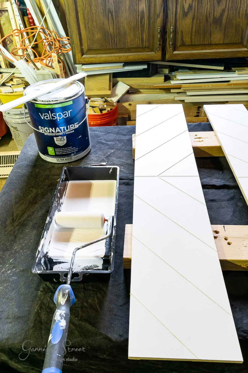 The workbench features chevron-grooved wooden boards. Nearby, a paint tray with a roller is partially covered in white paint from a can of Valspar Signature. In the background, cabinets and tools are visible.