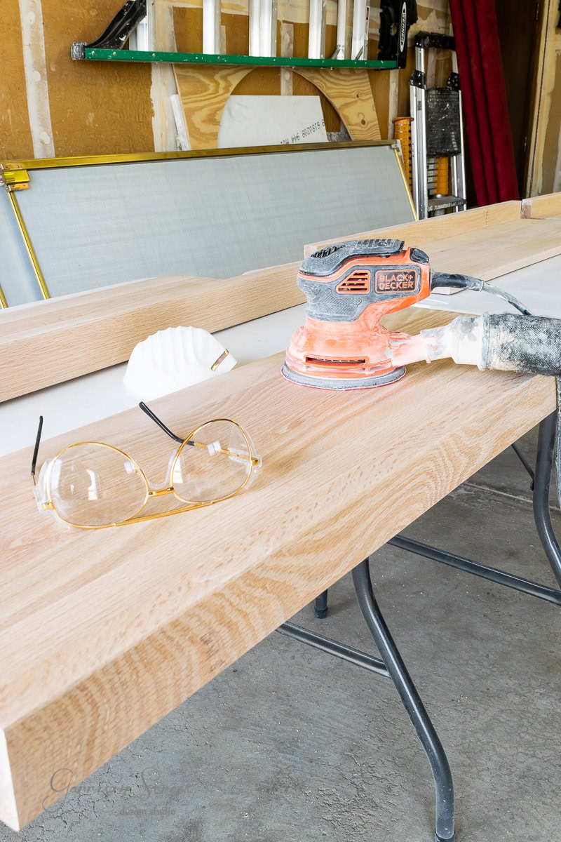 A wooden workbench with a sanding tool, a pair of clear glasses, and a white mask rests on it. In the background, tools and materials are leaning against the wall in a garage or workshop setting.