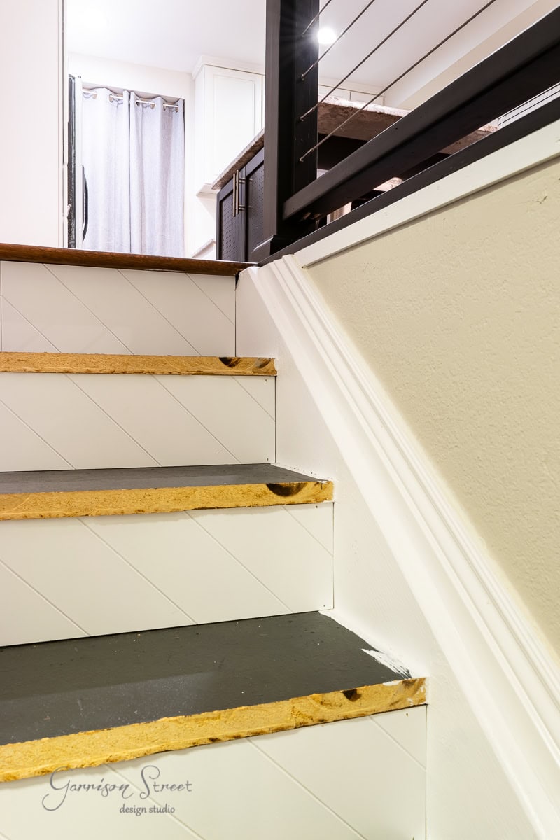A close-up view of indoor stairs mid-renovation showcases the DIY process: visible wood edges are partially finished, with some areas lacking trim and paint. A metal railing and part of the upper floor can be seen in the background.