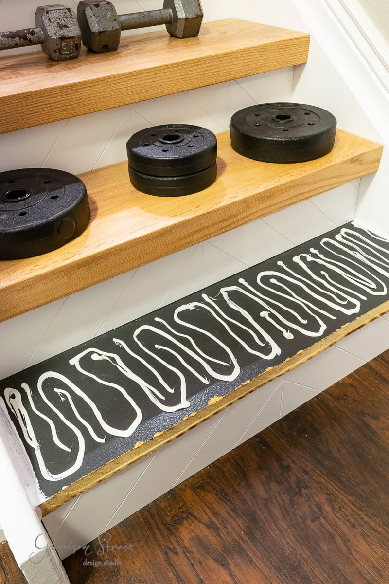 Wooden shelves holding dumbbells and weight plates are mounted on a wall above a hardwood floor. Decorative text in a white and black pattern is painted on the edge of the bottom shelf.
