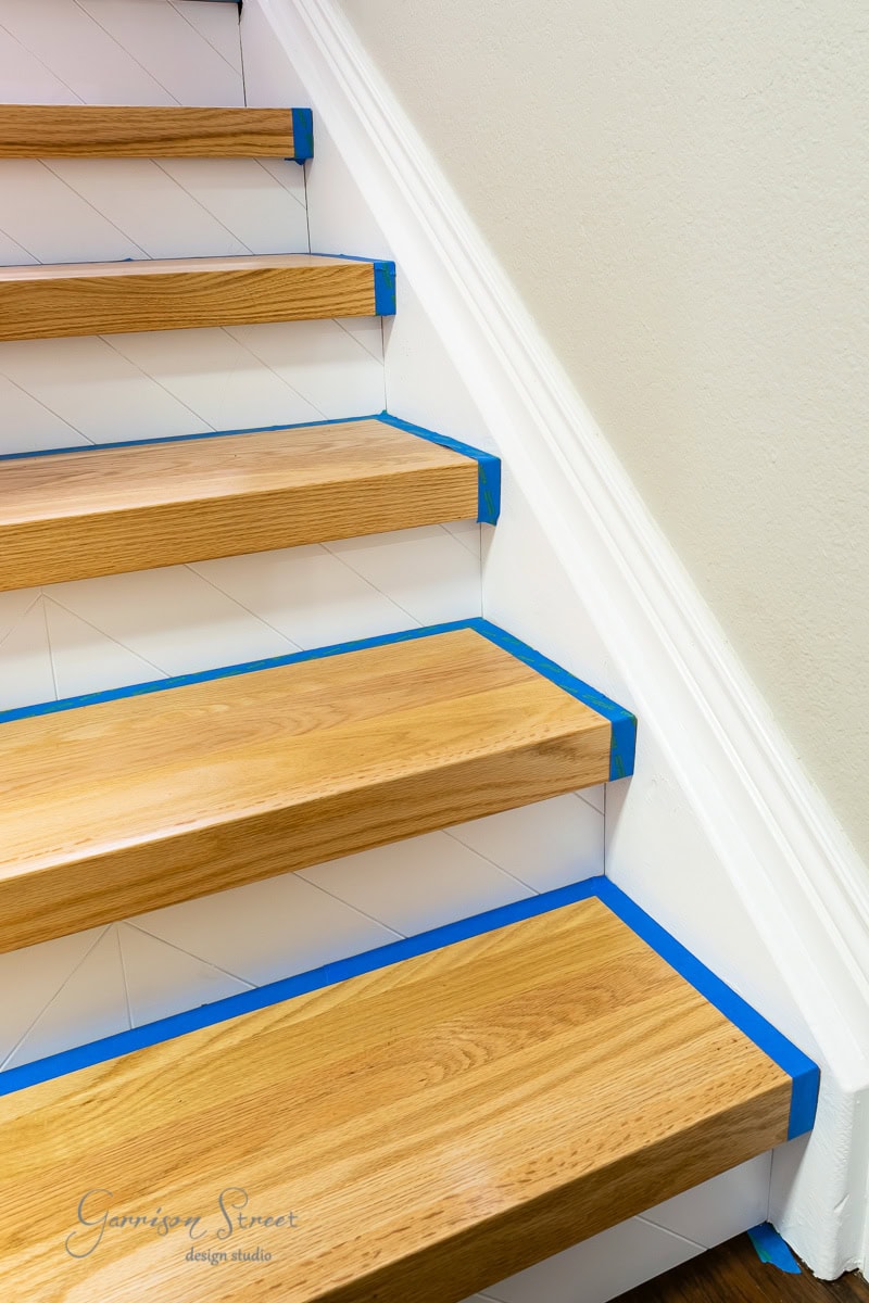 A staircase with wooden steps and white risers. Blue painter's tape lines the edges. The adjacent wall is a light cream color.