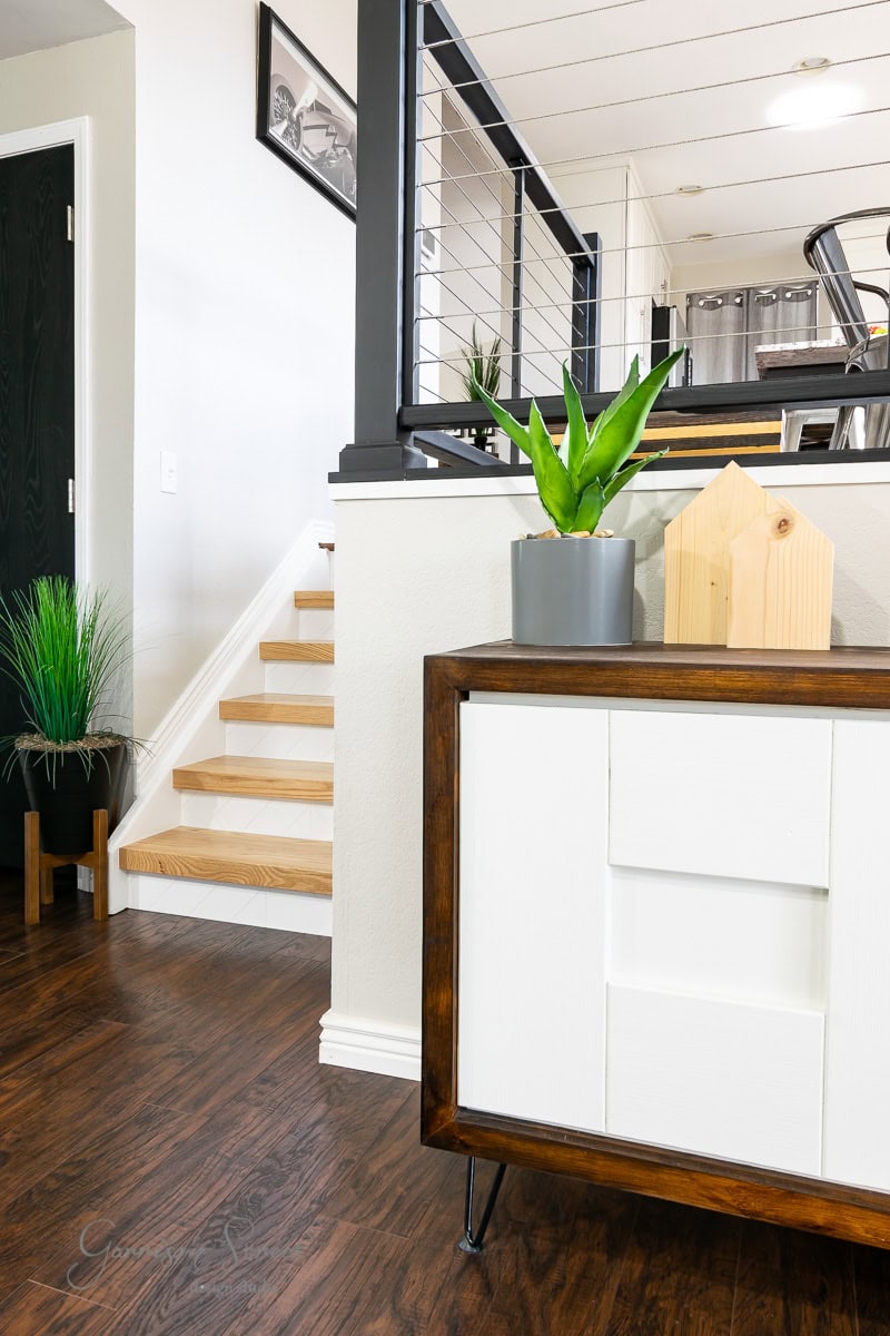 A modern interior showcases a wooden sideboard adorned with a plant and decor. The staircase features wooden steps leading to a sleek railing. Dark wooden floors complement the space, while a potted plant adds greenery on the left.