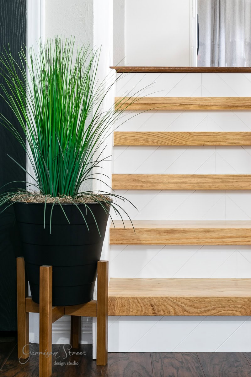 A potted green plant in a black pot with wooden legs stands beside a staircase adorned with wooden steps and white vertical risers.