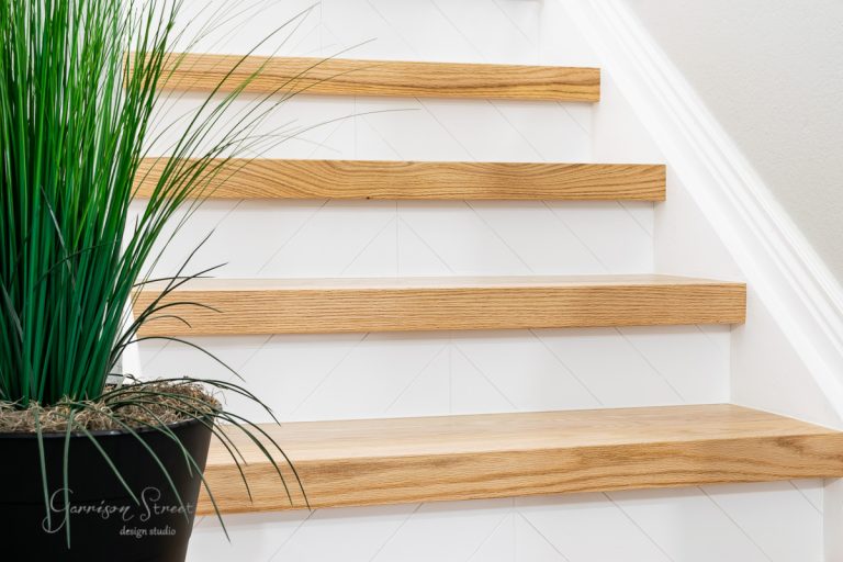 Discover how to remodel stairs DIY with a touch of elegance: this wooden staircase features light wood treads and white risers, enhanced by a chevron-pattern detail. A sleek black pot with tall, green grass-style plants adds a refreshing accent to the natural finish.