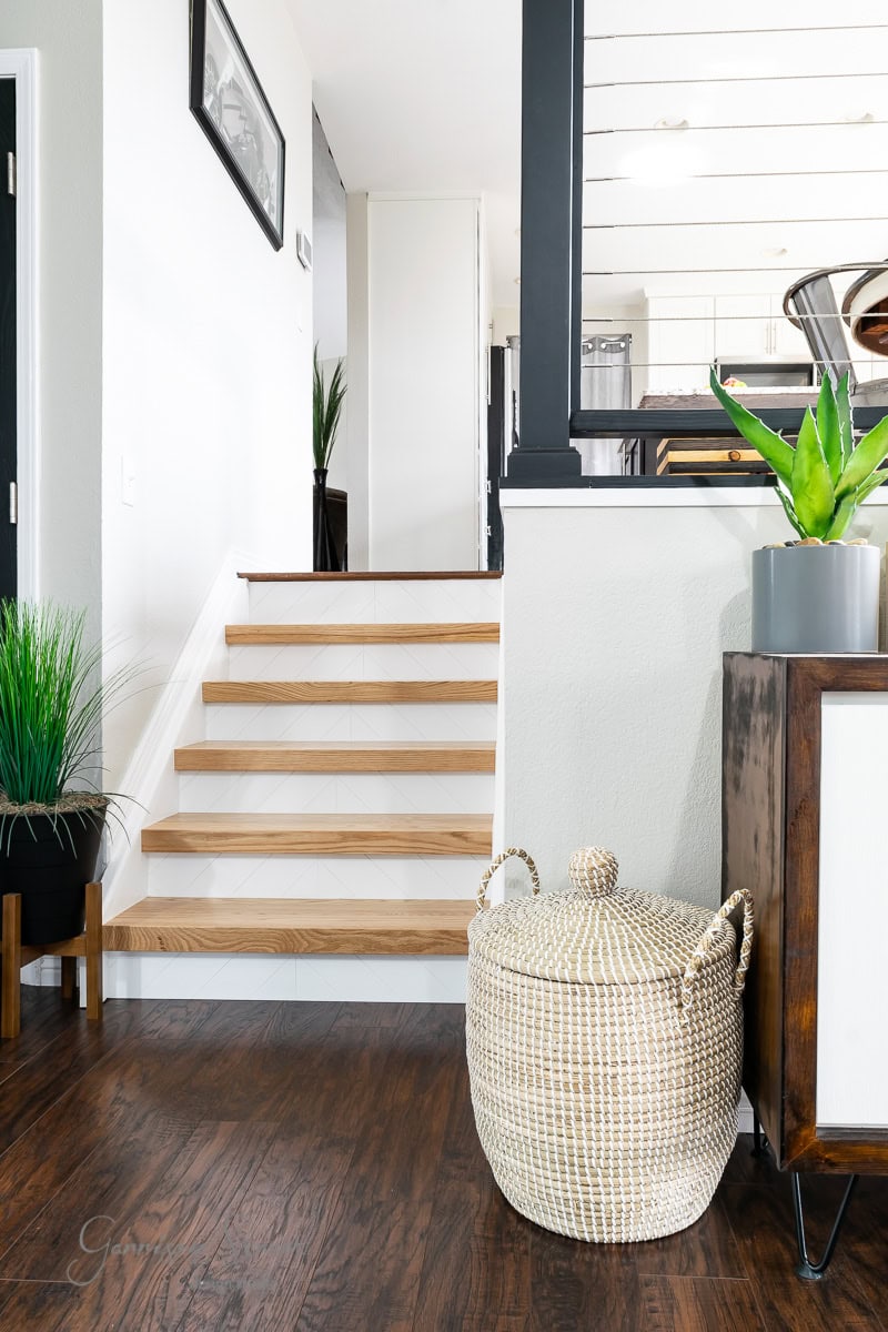 A modern interior showcases wooden stairs. A large woven basket sits on the dark hardwood floor next to a small cabinet, with potted plants artfully arranged for decoration. The kitchen is visible in the background.
