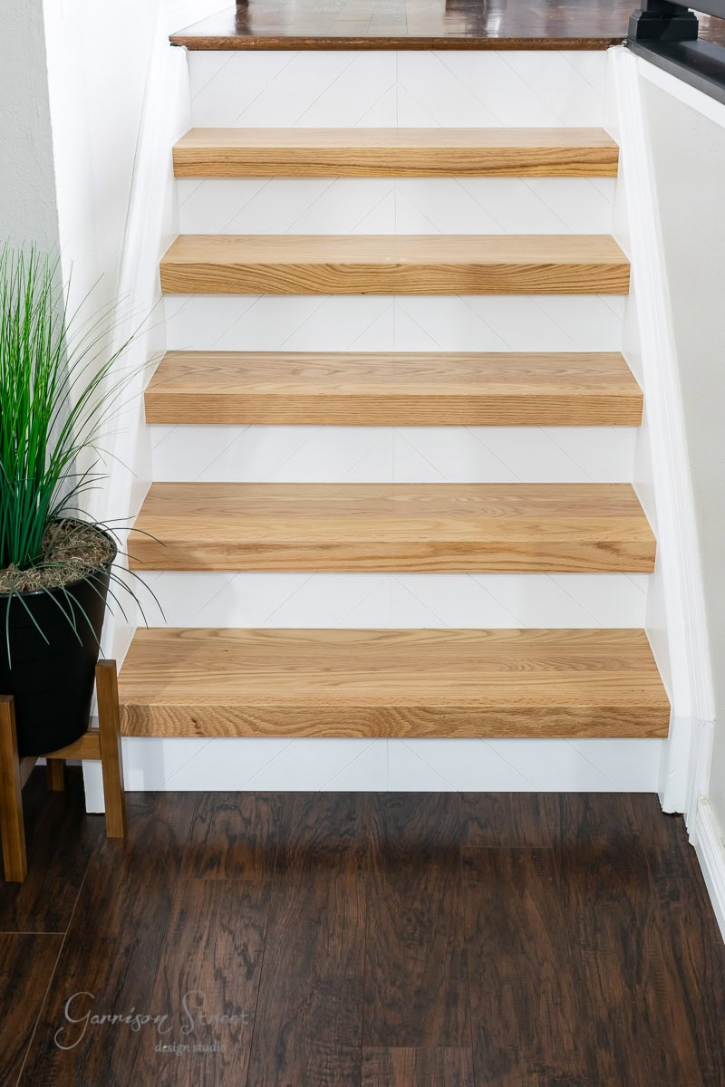 A staircase with five wooden steps showcases a modern design against a white backdrop. This setup features a wooden floor at the base, complemented by a potted plant with green foliage in the bottom left corner.