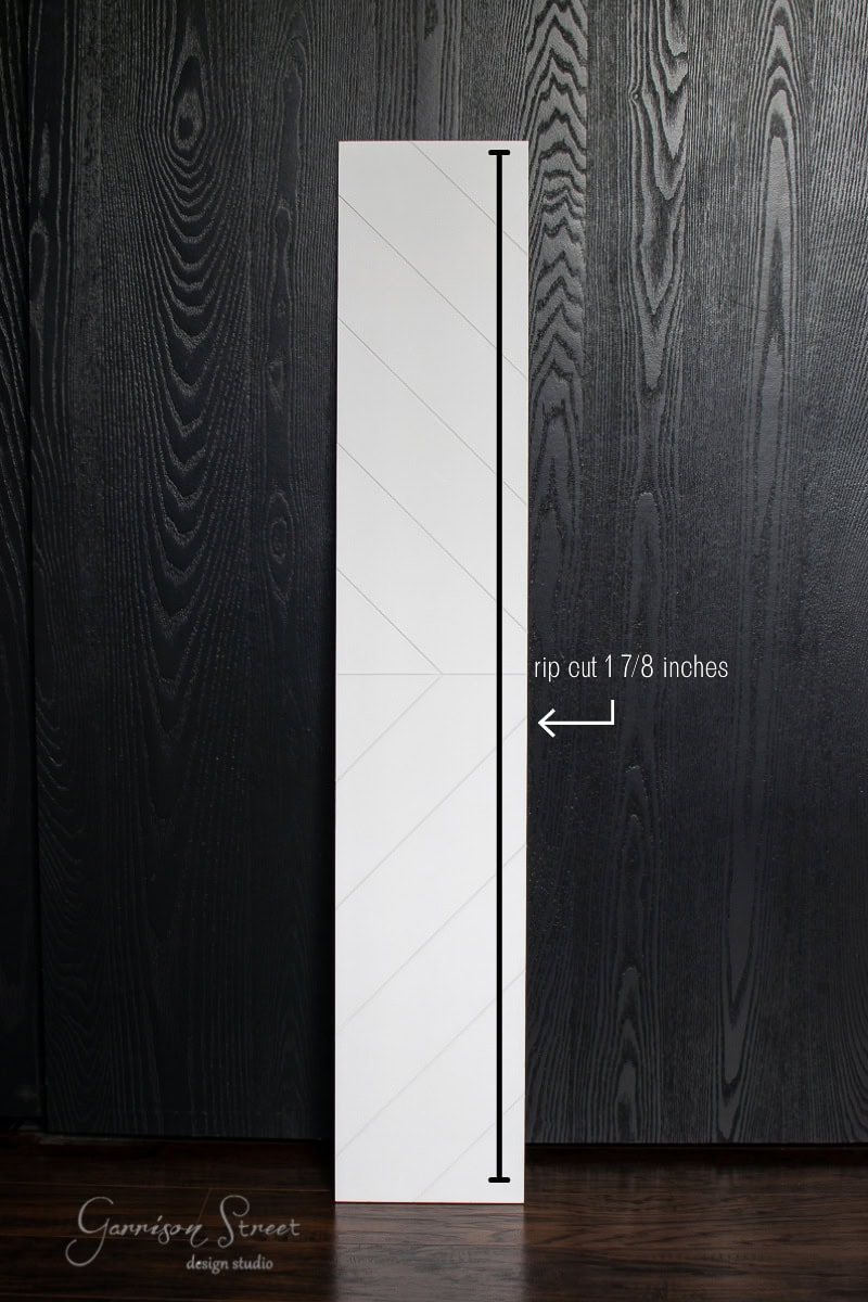 A white wooden plank with a faint diagonal pattern stands vertically against a dark textured wall. An arrow highlights a rip cut measurement of 1 7/8 inches on the plank.