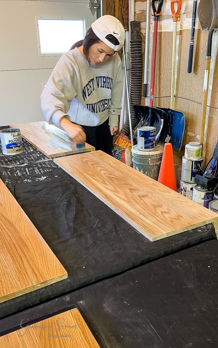A person wearing a West Virginia University sweatshirt and a cap applies a finish to wooden planks on a covered table in a workshop. Various tools and supplies are scattered in the background.