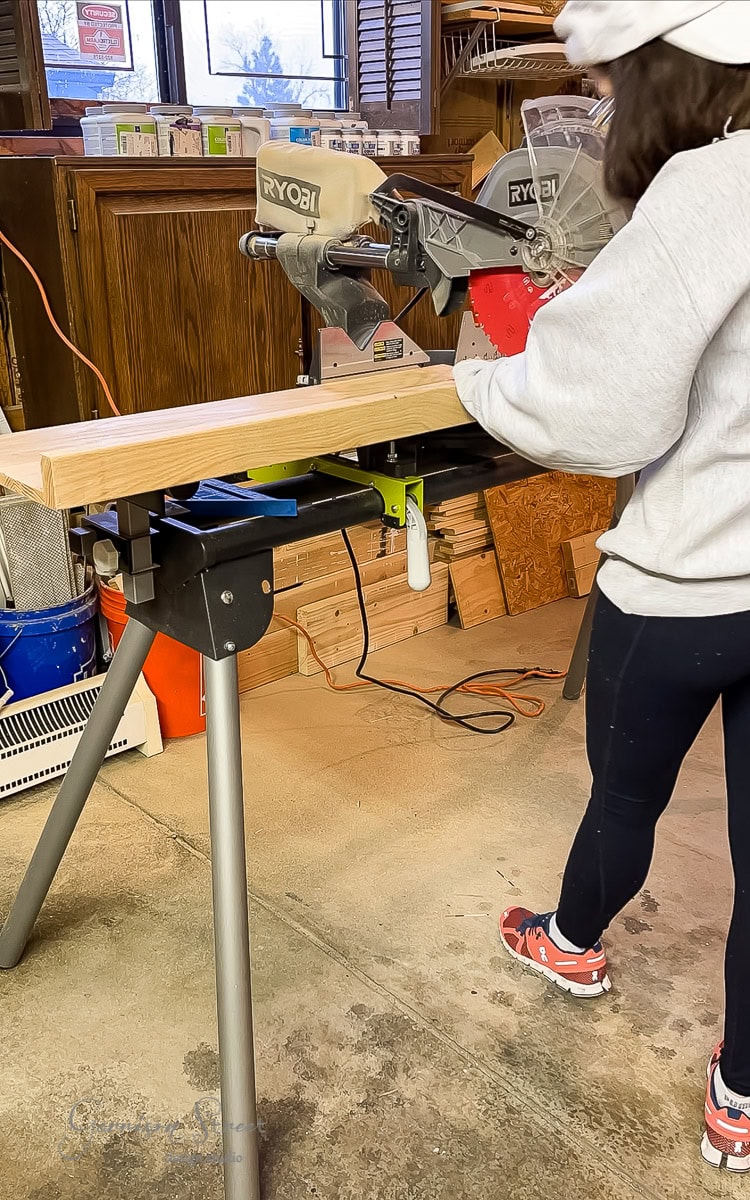 In a workshop filled with wooden planks and tools, a person in a white hoodie and red sneakers uses a Ryobi miter saw to cut a board.