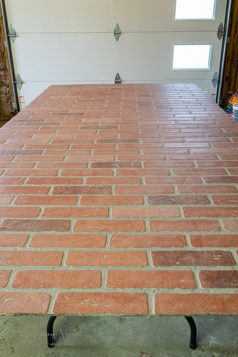 A large table with a surface resembling a brick pattern is set in a garage with a white garage door in the background, offering unique basement wall ideas not drywall. The table's legs are black, complementing the bricks' various shades of red and brown.