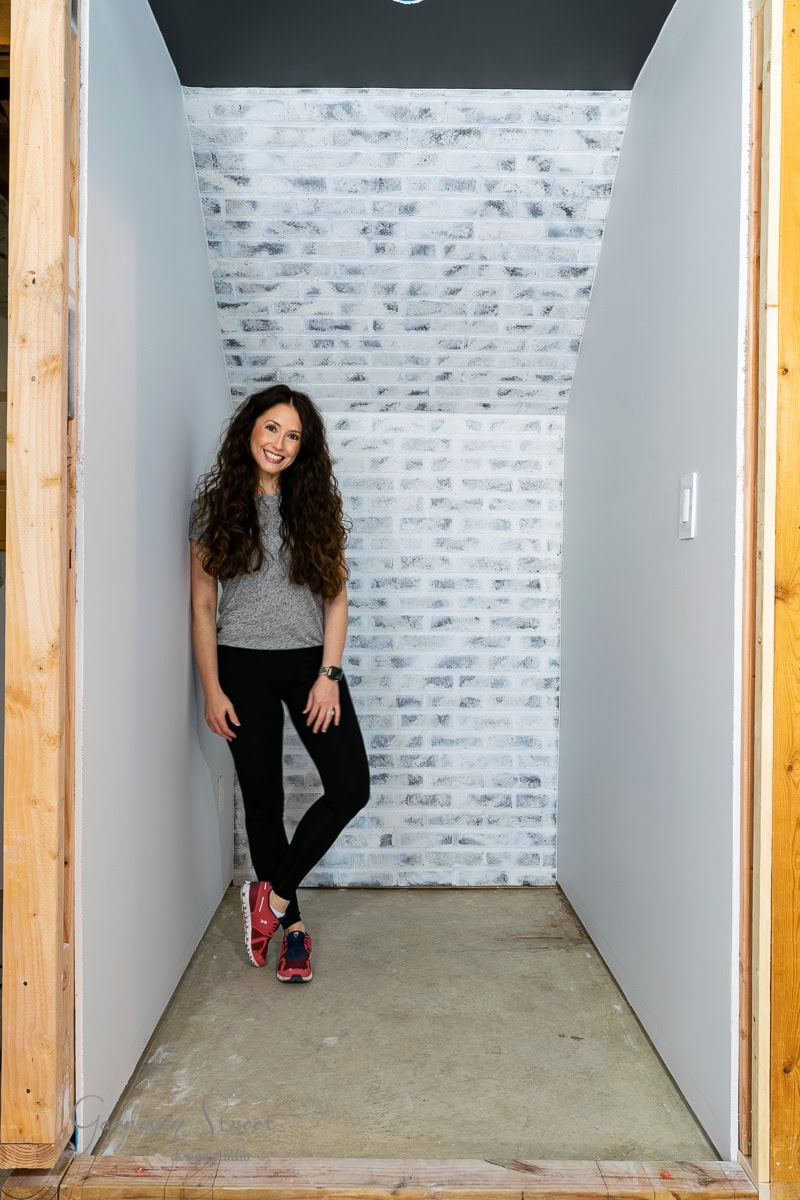 A person with long curly hair, wearing a gray top, black pants, and red sneakers, stands smiling against a white brick wall in a narrow, unfinished space showcasing basement wall ideas not drywall with wooden framing and an exposed concrete floor.
