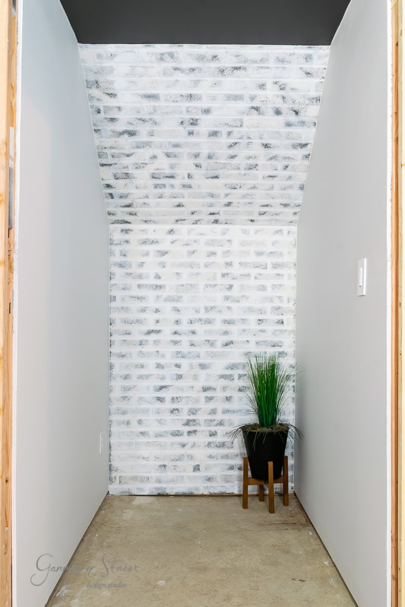 A narrow, empty hallway with white brick walls and a slanted ceiling. Embracing basement wall ideas not drywall, a potted plant with tall, grass-like leaves sits on a wooden stool against the brick wall. The floor remains plain and unfinished.