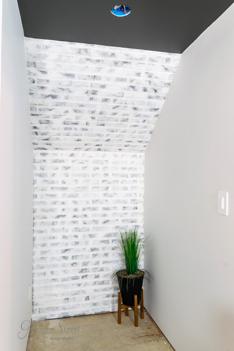 A small nook showcases basement wall ideas with a whitewashed brick wall and a gray ceiling. A black planter filled with green grass nestles on a wooden stool in the corner. The floor is bare concrete, and an unlit circular light fixture is embedded overhead, avoiding traditional drywall designs.