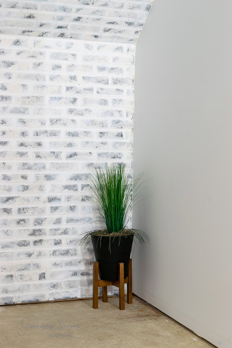A tall plant in a black pot stands on a wooden stand against a whitewashed brick wall, showcasing basement wall ideas beyond drywall. The light beige tile floor complements the minimalist decor in the corner of a plain gray wall.