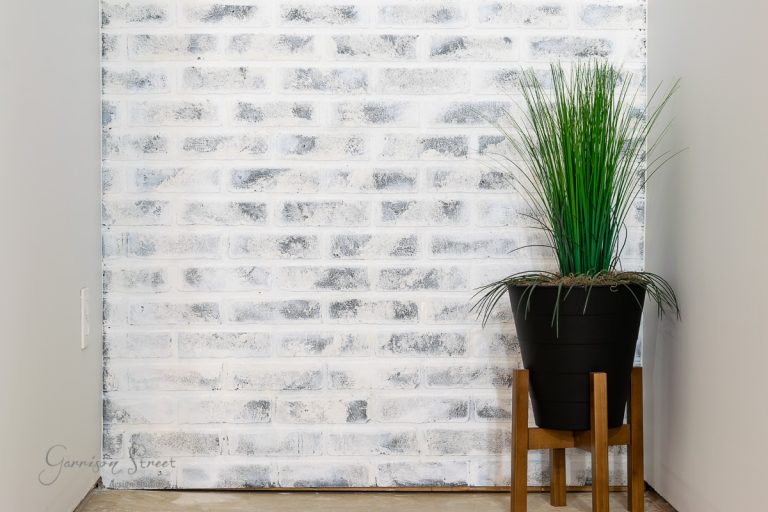 A green plant in a black pot sits on a wooden stand against a white brick wall, offering inspiration for basement wall ideas beyond drywall.