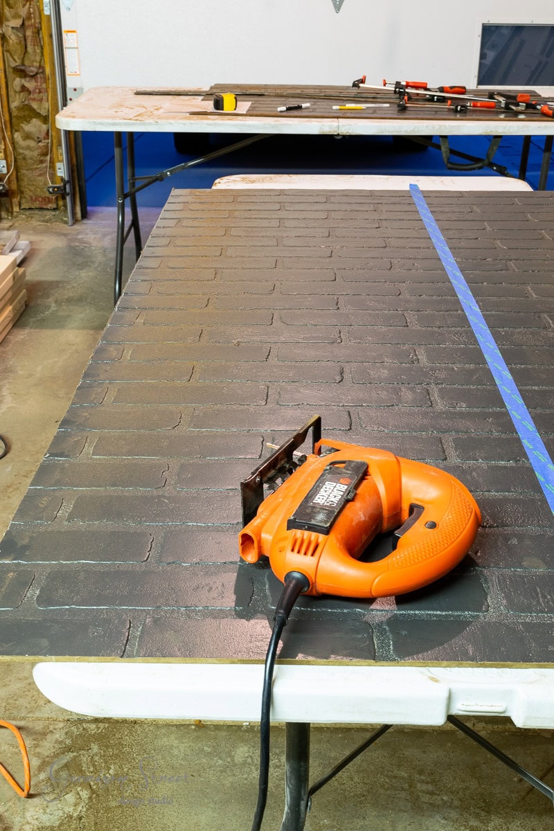 An orange power jigsaw rests on a table with a faux brick panel, ideal for basement wall ideas not drywall. A measuring tape and clamps are visible in the background, completing the workshop or construction area scene.