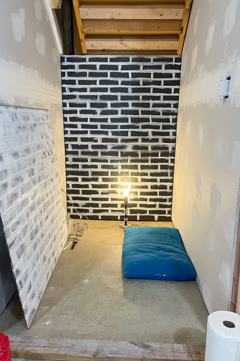 A small, unfinished basement nook features a black and white brick pattern wall, offering unique basement wall ideas not drywall. A blue cushion rests on the floor beneath a standing lamp, with a partially painted brick panel leaning against the left wall.