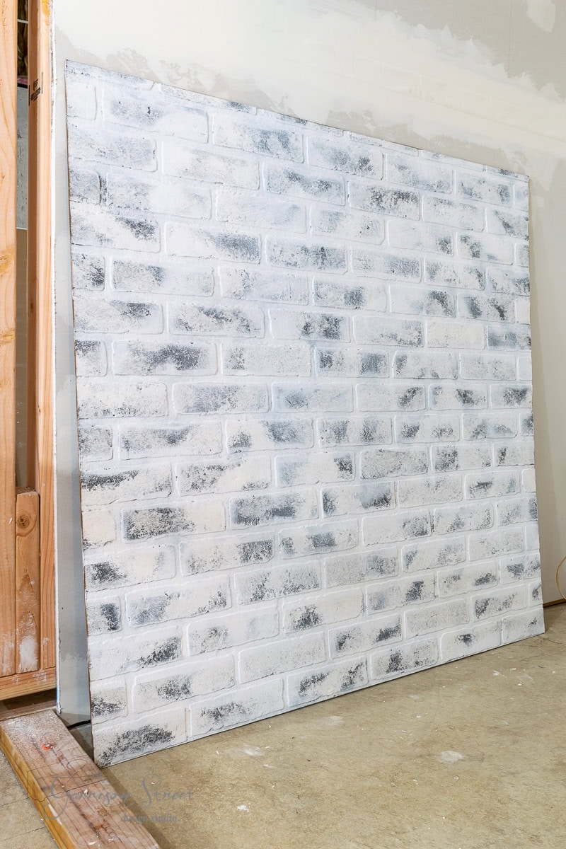 A large panel painted to resemble a whitewashed brick wall leans against a partially constructed wooden frame and an unfinished wall in a room with a concrete floor, offering creative basement wall ideas that go beyond traditional drywall solutions.