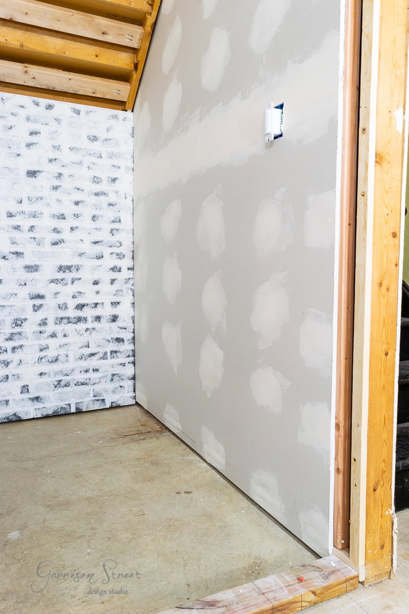 A partially finished interior wall showcases alternative basement wall ideas, with white patches over seams and screws. The concrete floor is visible, and a partially painted brick wall is on the left. Wooden beams are exposed on the ceiling, suggesting a move beyond traditional drywall.