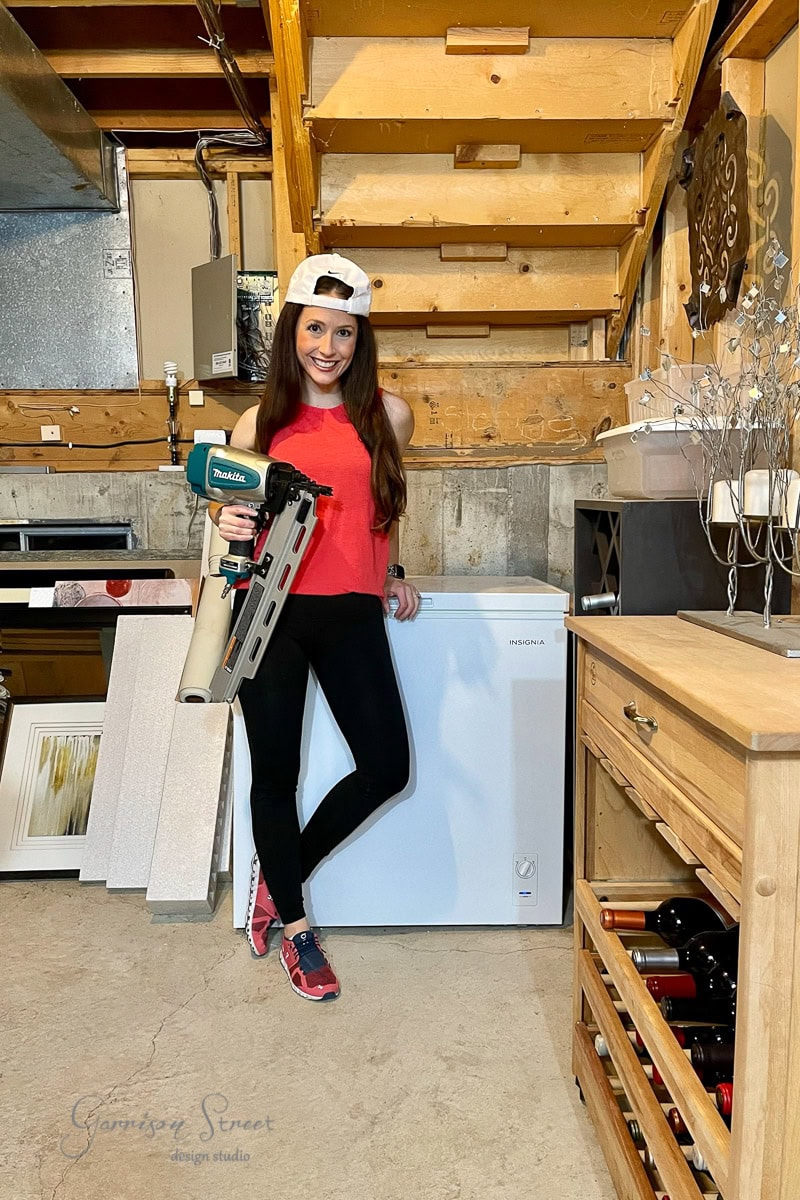 A woman in a red tank top and black pants stands in a wine cellar next to a freezer, holding a nail gun. She wears a backwards white cap and pink sneakers. The cellar has wooden shelves with various items stored around.