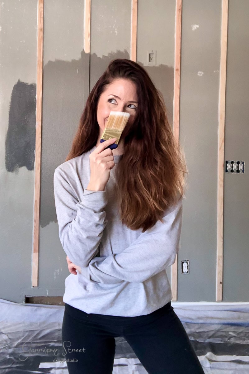 A woman with long brown hair playfully pretends to eat a paintbrush. She stands in a basement under renovation, with drywall and partially painted walls in the background. She wears a light gray sweatshirt and black pants.