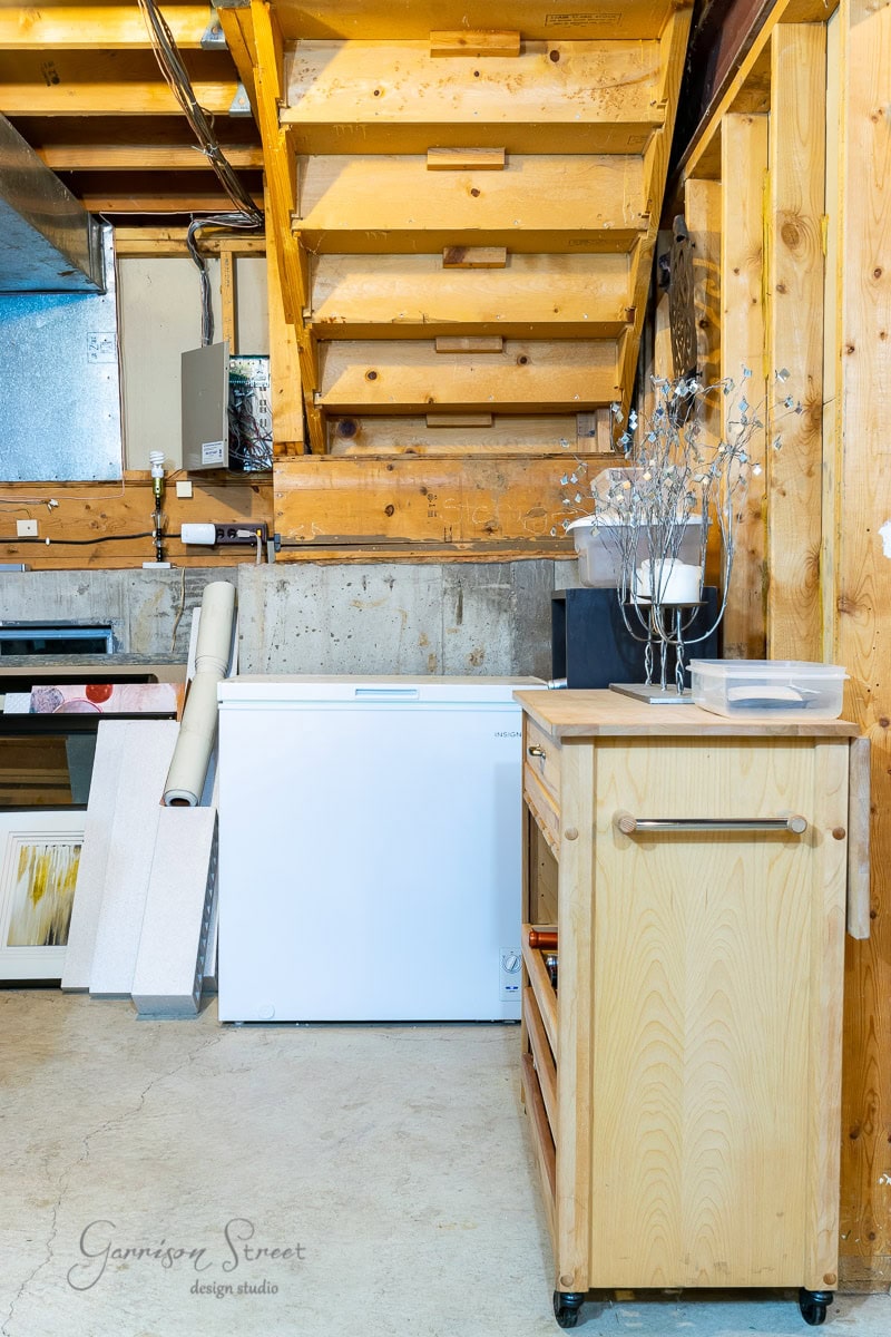 The basement, with its wooden stairs and white chest freezer, exudes a rustic charm. A light wooden cart complements the space, adorned with rolled-up paper and a tall vase of branches. It's as practical as it is inviting—perfect for transforming into a cozy wine cellar.