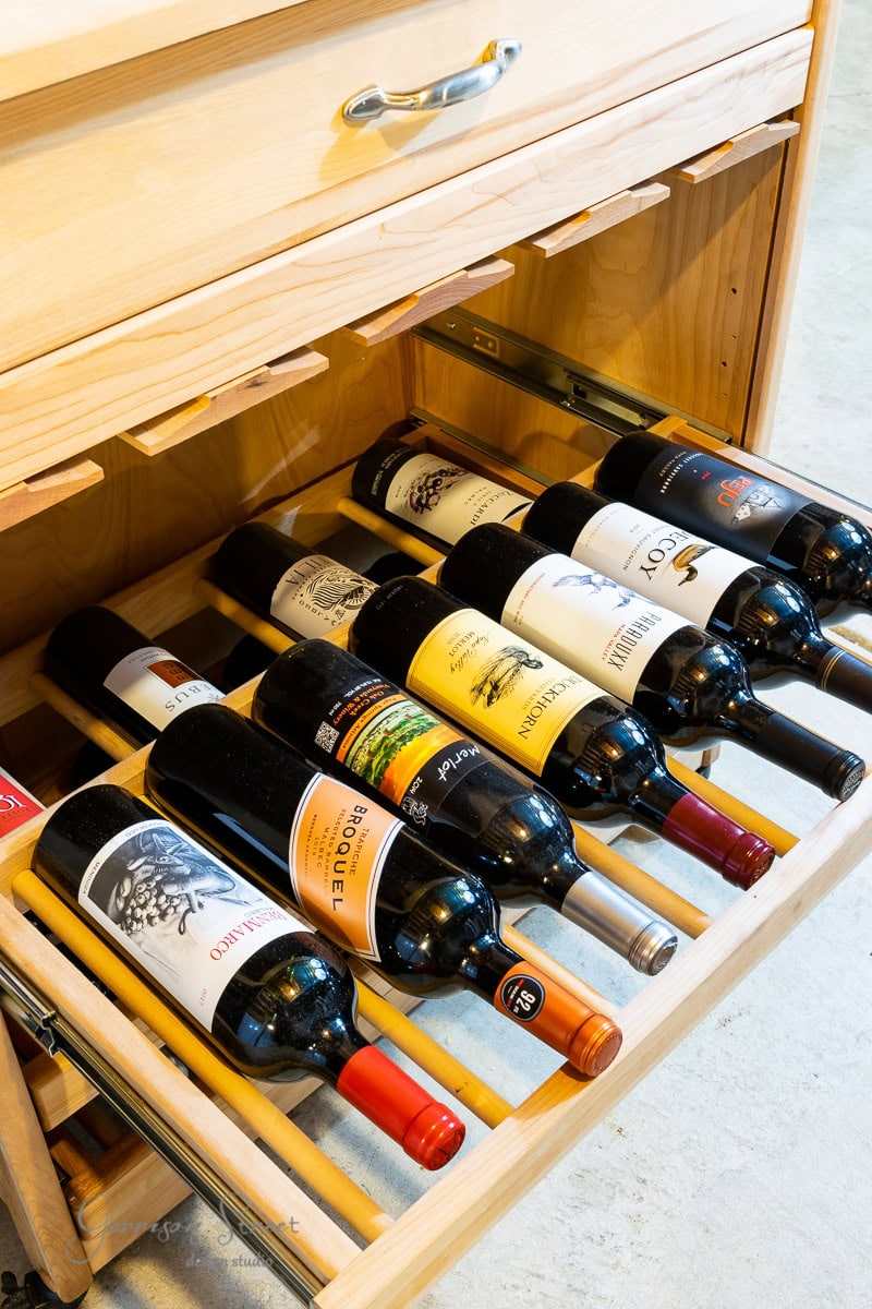 A wooden drawer is open, revealing nine bottles of wine neatly organized on racks. Each bottle boasts a unique label, nestled in the light-colored cabinet's basement wine cellar. The scene is well-lit, showcasing the curated collection.