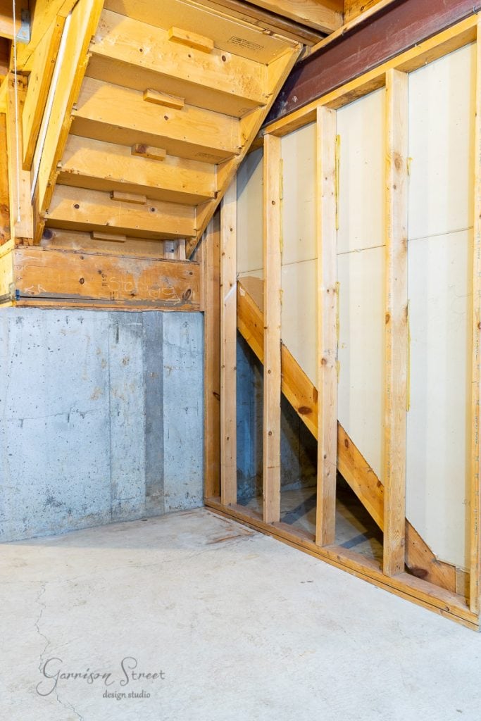A partially finished basement with exposed wooden framing and stairs offers potential for transformation into a cozy wine cellar. The concrete floor and walls contrast with the wood structure. "Garrison Street Design Studio" is written in the lower left corner.