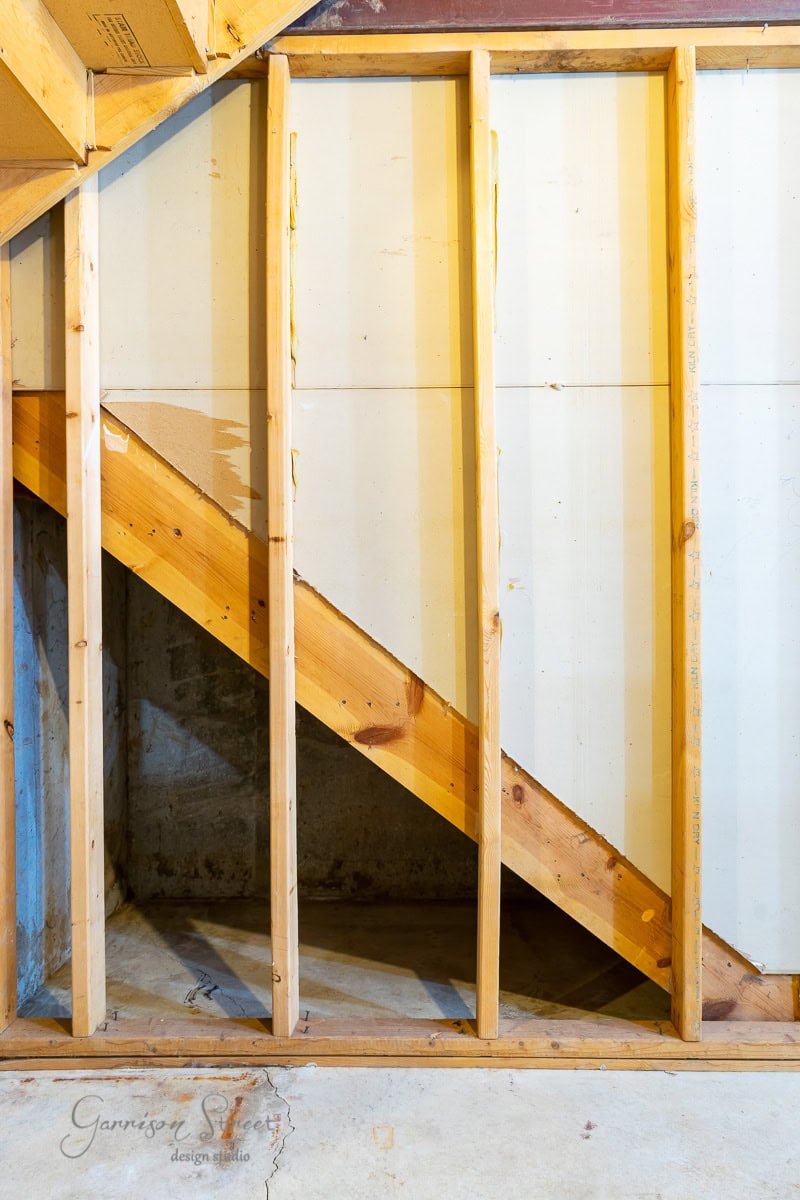 Framed section of a basement under construction, showing wooden beams and a diagonal support beneath stairs, hinting at the future potential for a cozy wine cellar amidst the concrete and panel wall.
