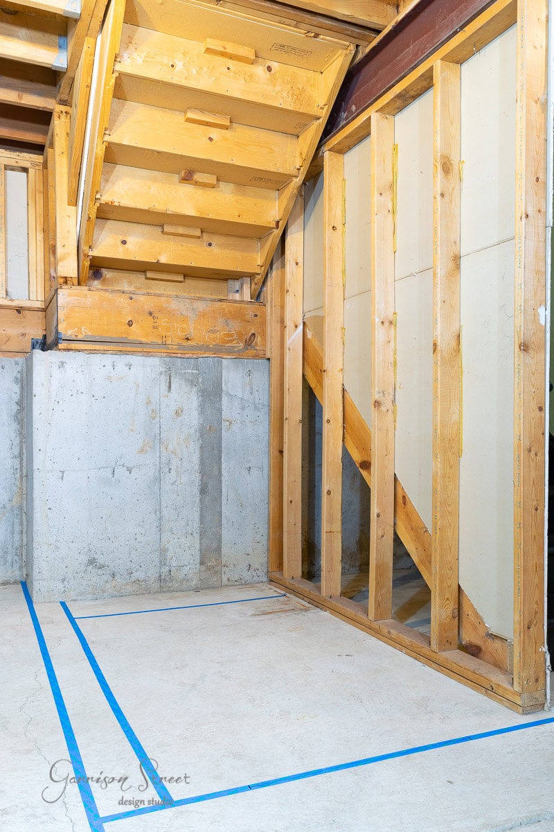 A basement under construction with exposed wooden framing and concrete walls shows how to frame basement walls. The ceiling is an unfinished wooden staircase, while blue tape outlines sections on the concrete floor.