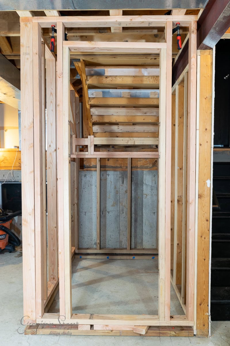 The wooden framework of a small under-staircase storage room is under construction, showcasing an open doorway and stairs without steps. The space illustrates how to frame basement walls, with visible wood beams and concrete walls surrounding the area.